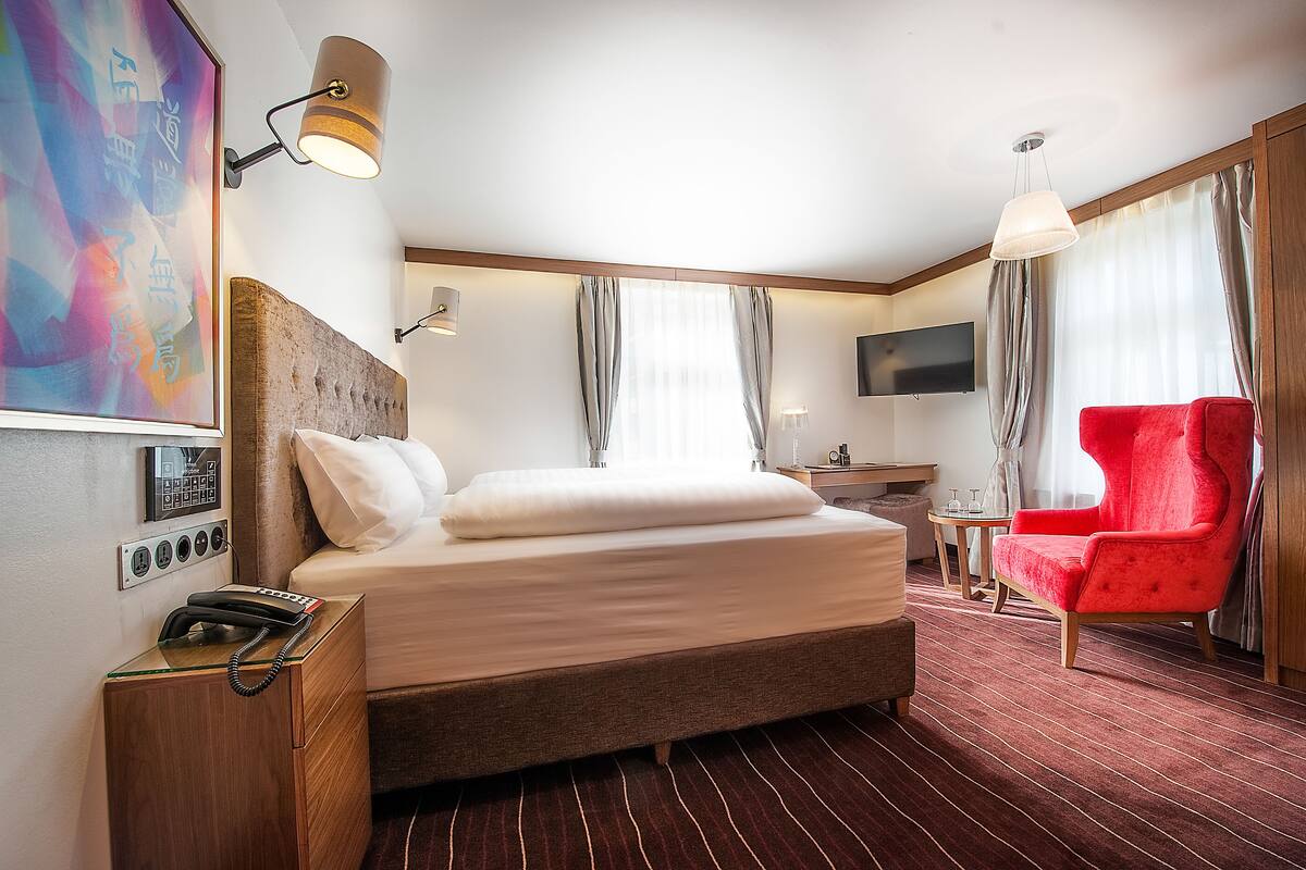 Habitación de hotel con una cama grande, un sillón rojo, un escritorio con una silla, un televisor y un teléfono en la mesita de noche.