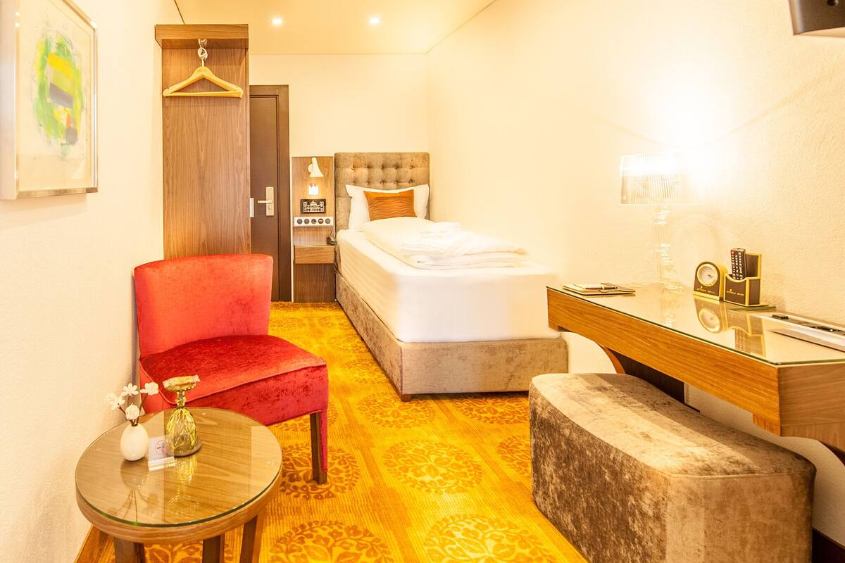 Habitación de hotel con una cama, una silla roja, una mesita, un espejo y una mesita de noche con una lámpara.