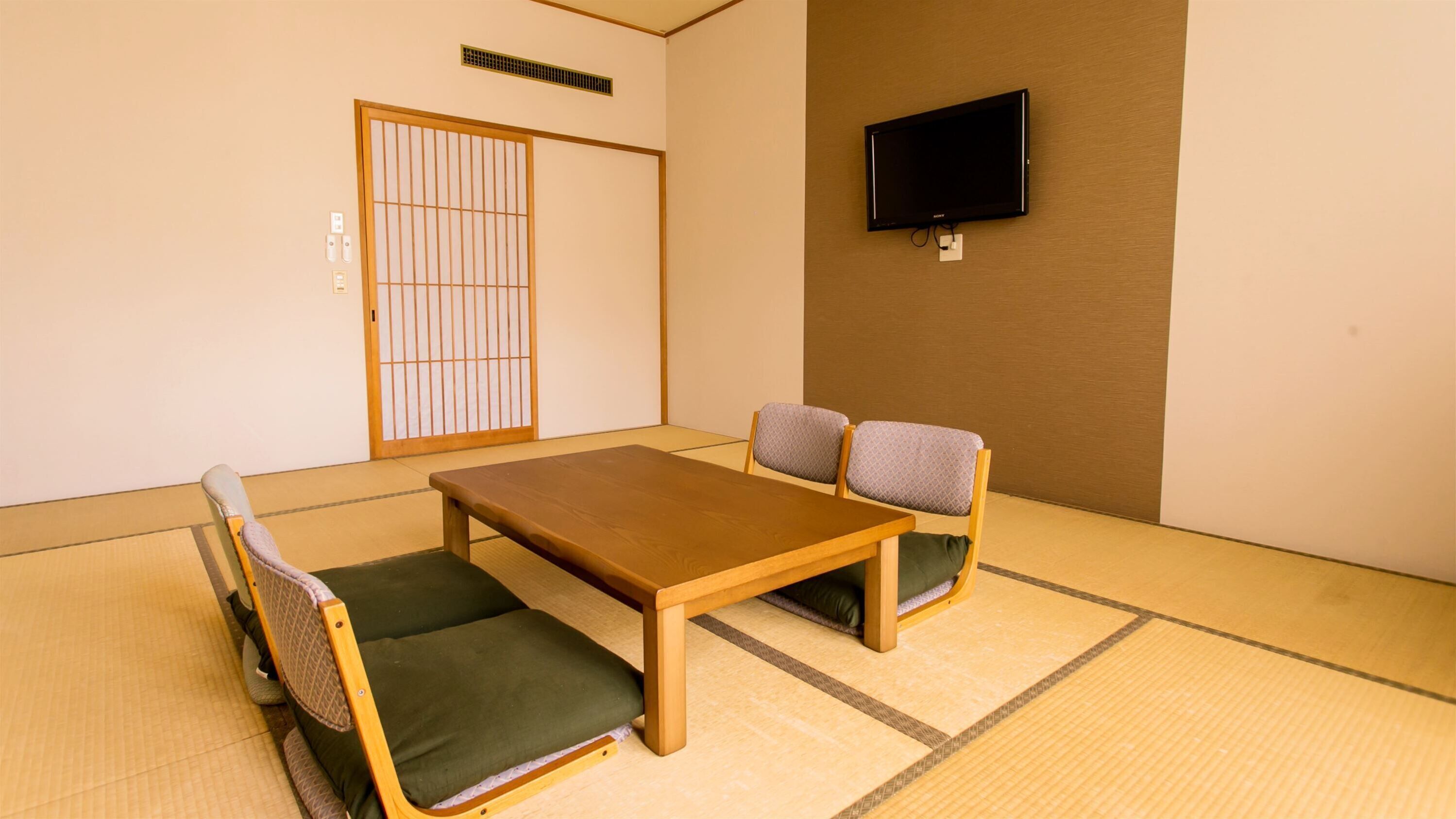 A traditional Japanese room with tatami flooring, a low wooden table, two cushioned chairs, a sliding shoji screen door, and a mounted television.