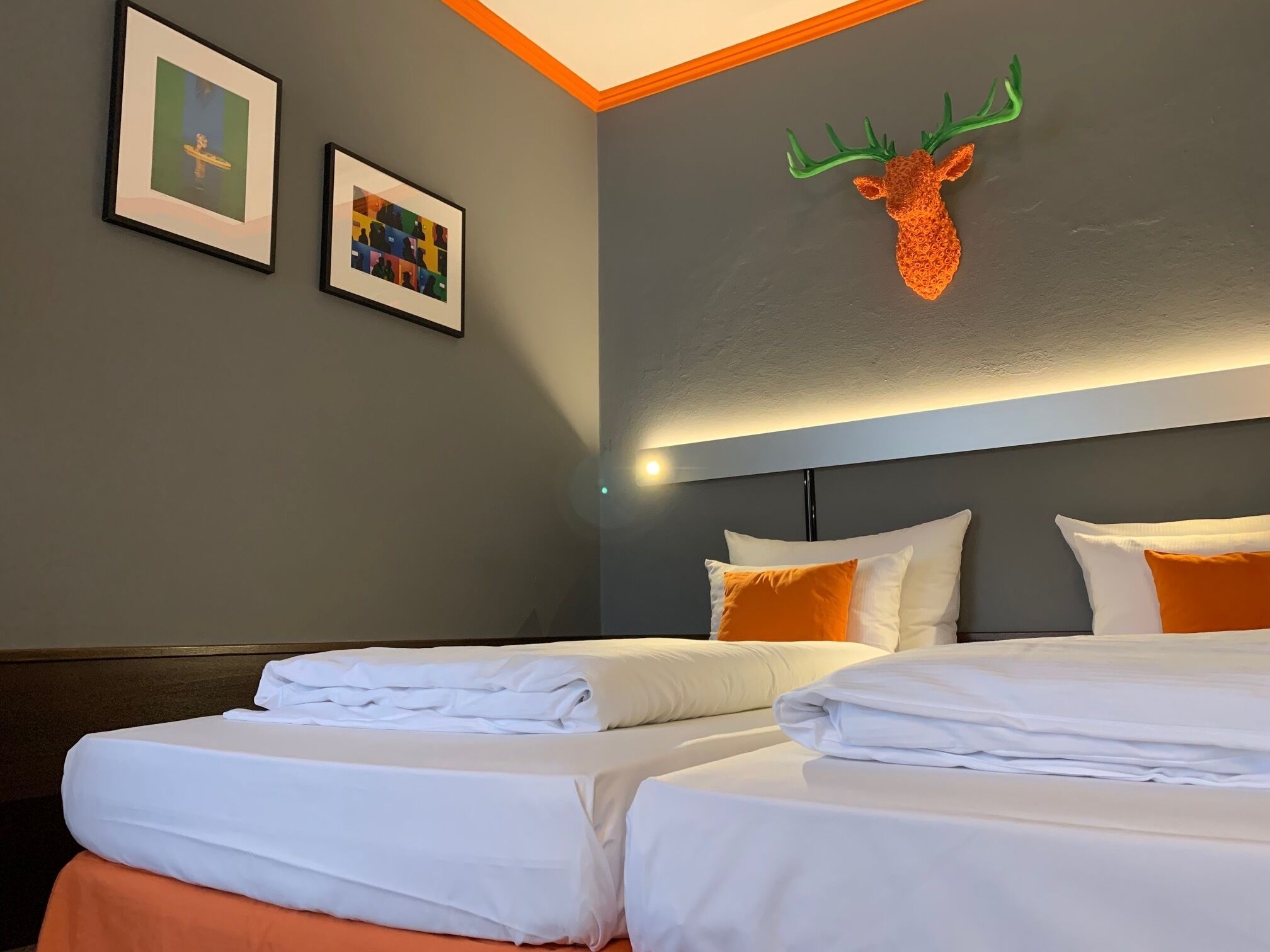 A hotel room with two beds, white bedding, a headboard with orange accents, and two framed artworks on the wall.