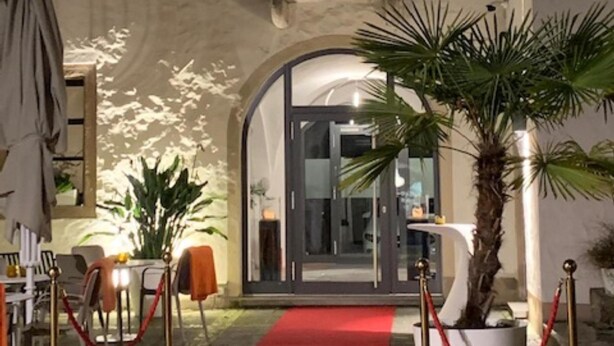 An entrance with a red carpet, a palm tree, and a building with arched windows. - Hotel Maxplatz (Hof)