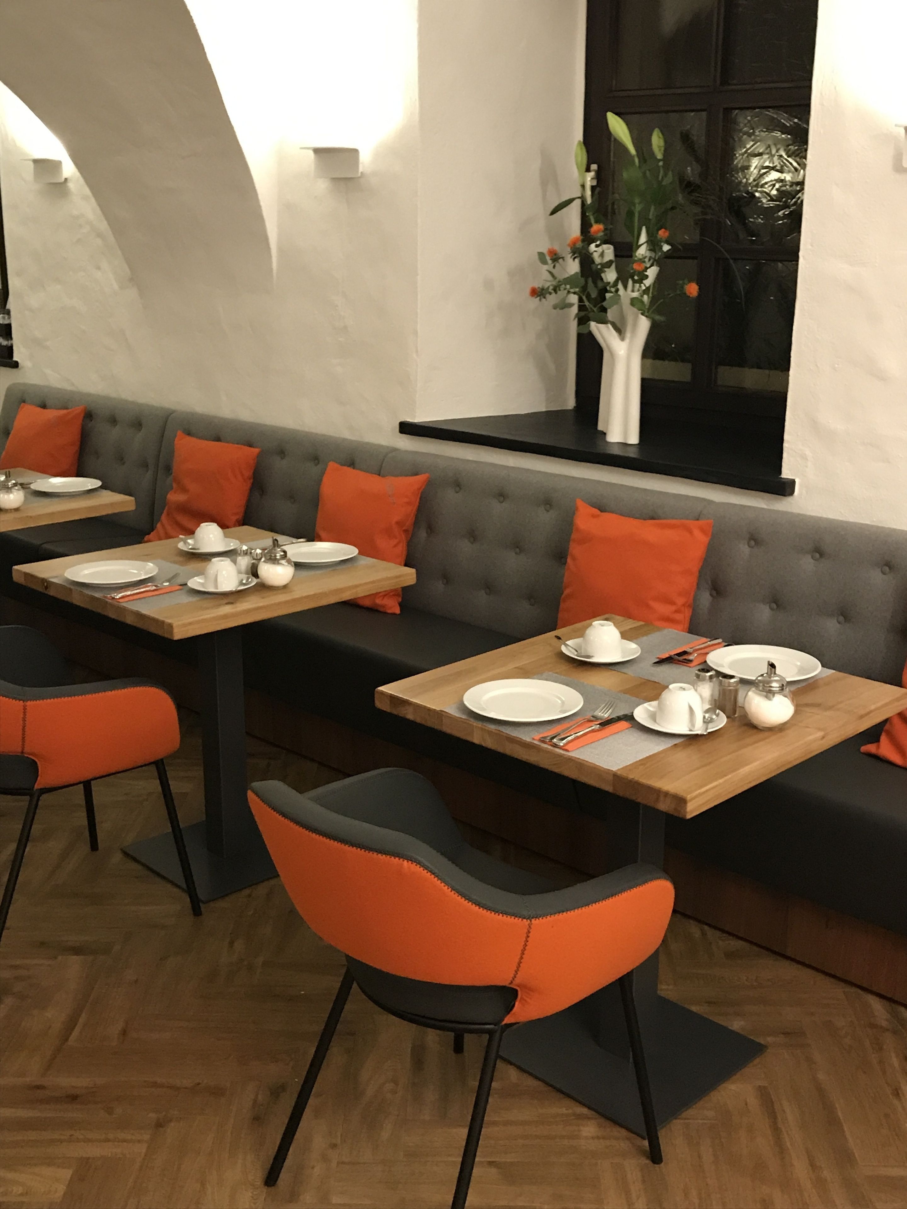A restaurant with a modern interior, featuring a long grey sofa with orange cushions, wooden tables, and black and orange chairs.