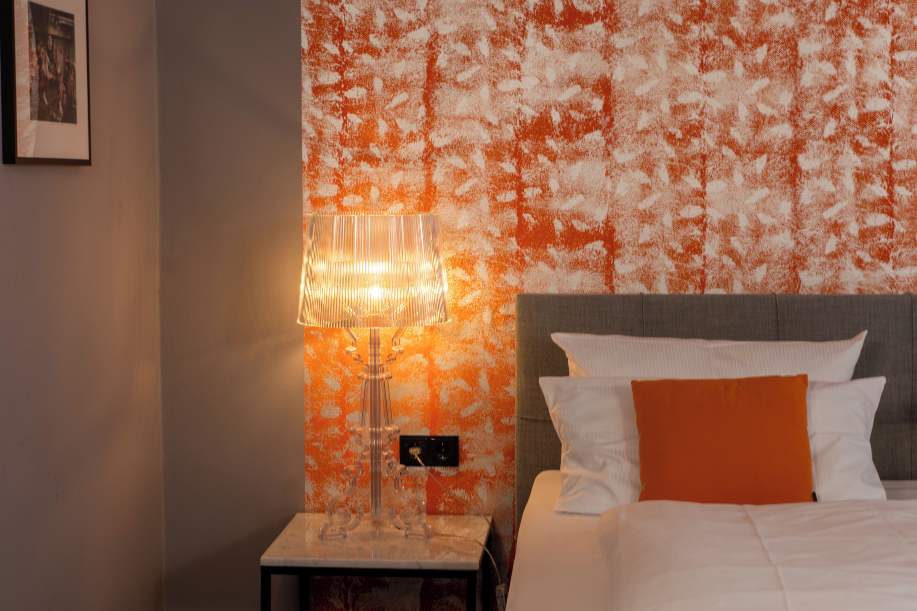 A hotel room with a bed, a nightstand, a lamp, and a textured orange wall.