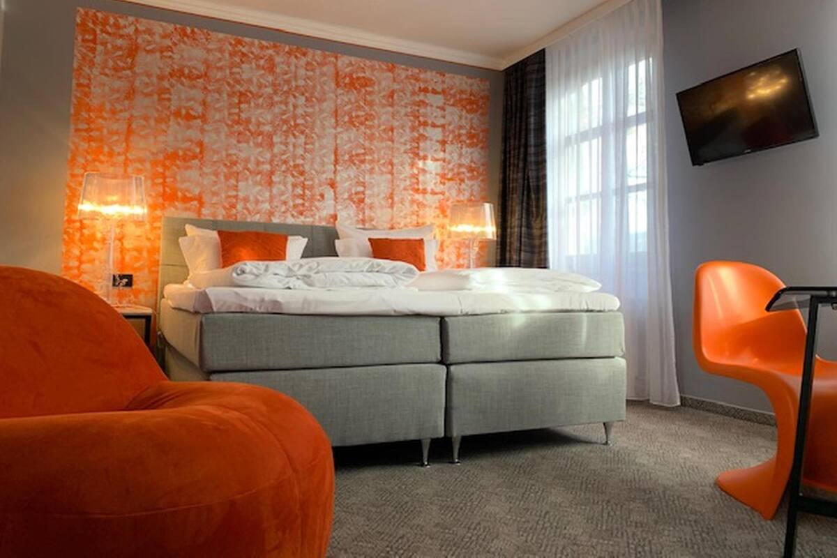 A modern hotel room with a grey bed, orange accent chairs, a flat-screen TV, and a patterned orange wall.