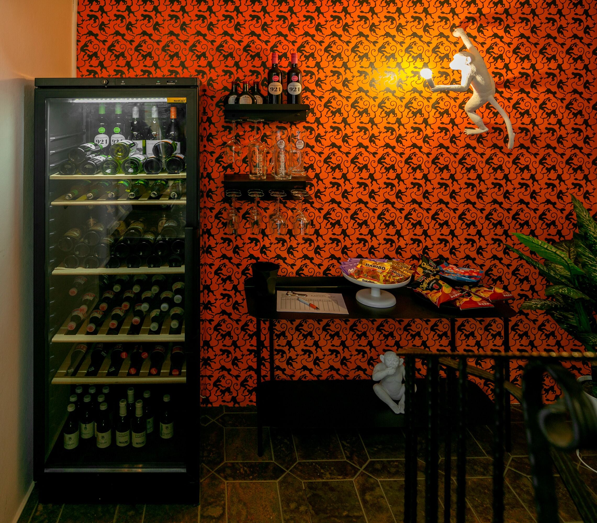 A bar area with a refrigerator stocked with bottles, a shelf with glasses, and a monkey statue.