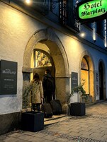 Entrada de un hotel con un arco, un letrero que dice 'Hotel Maxplatz' y un cartel de 'HAAR-KUNST' al fondo.