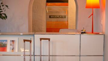 A hotel reception desk with a sign reading "MAXPLATZ," a lamp, and a suitcase.
