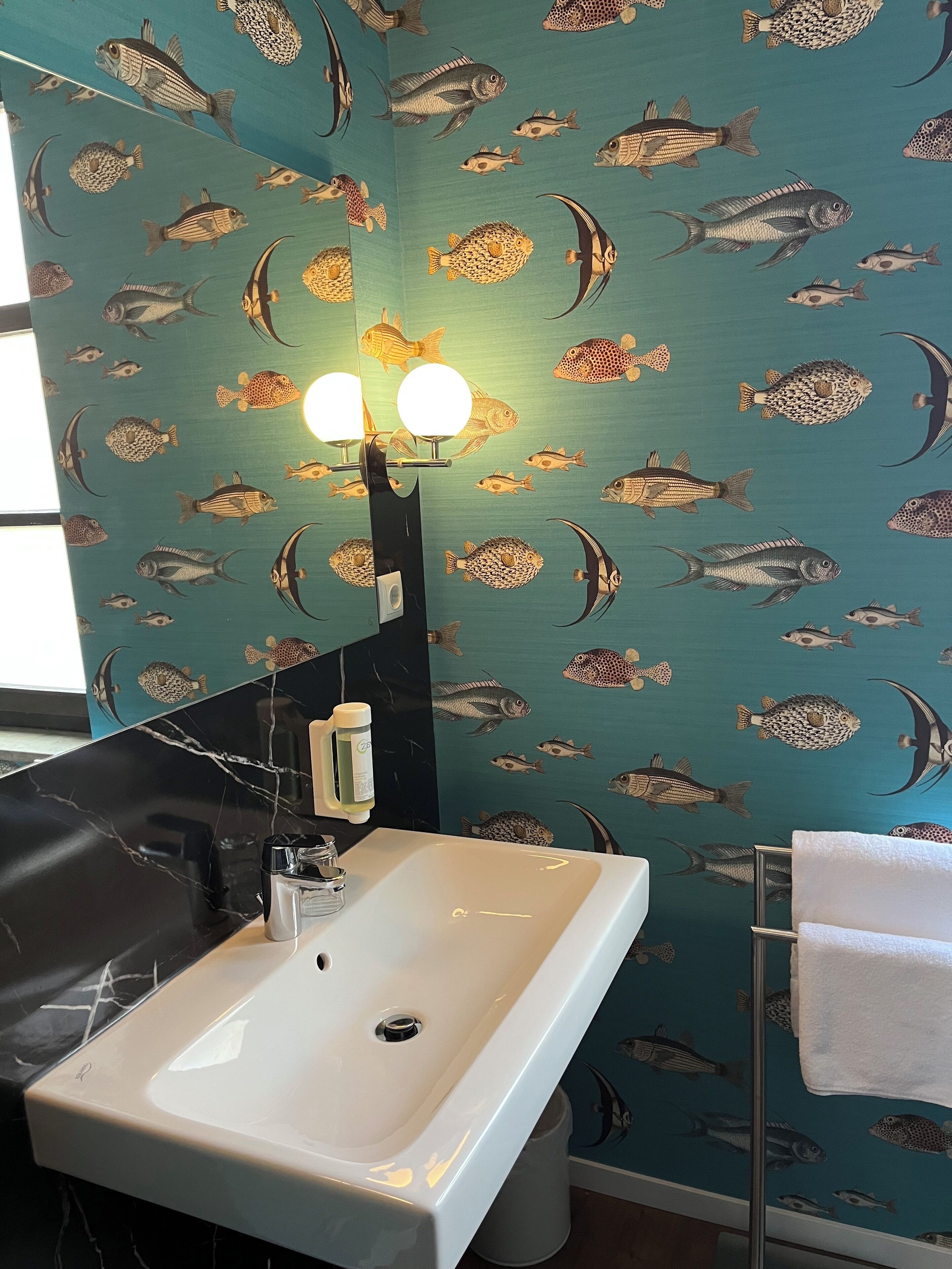 Ein Badezimmer mit Waschbecken, Spiegel und Handtuchhalter. Die Wand zeigt ein Fischmuster.