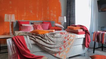 A modern bedroom with a grey sofa, a bed with orange and grey bedding, and a TV on the wall.