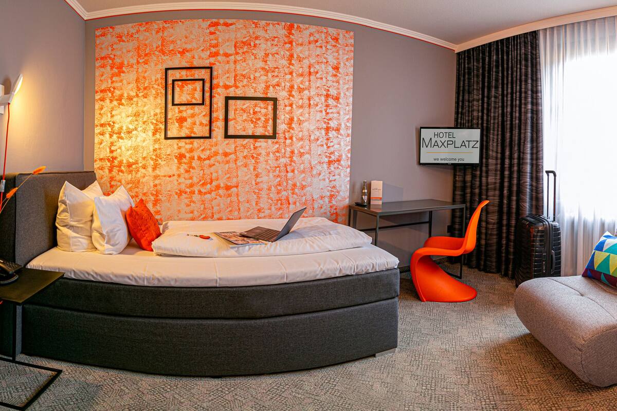 A modern hotel room with a curved bed, a desk, and a comfortable armchair.