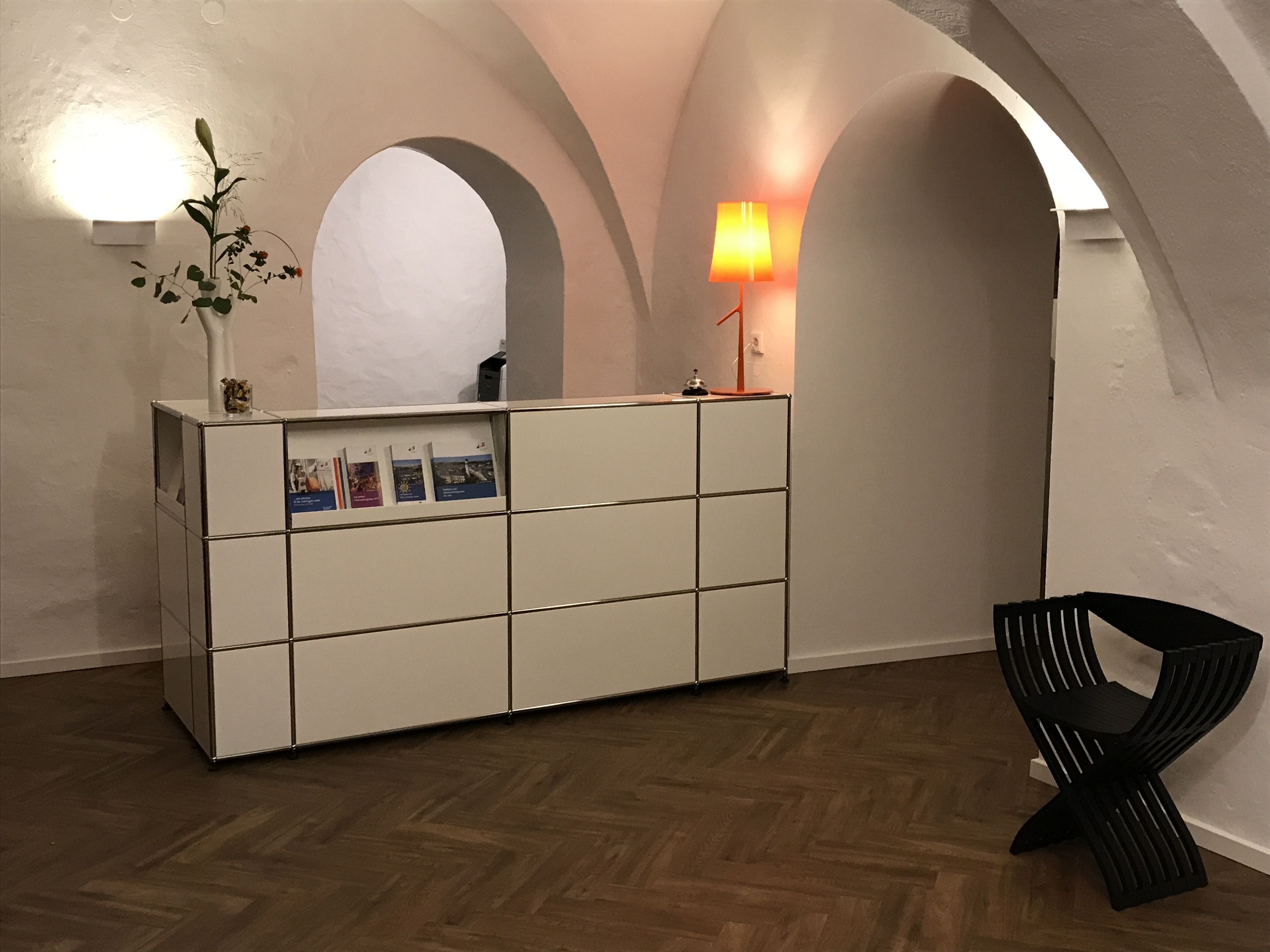 Un mostrador de recepción con una planta, una lámpara y una silla en una habitación con techos abovedados y suelo de madera.