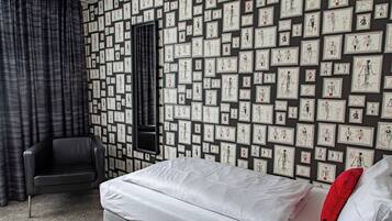 A hotel room with a bed, a chair, a large window, and a wall covered with framed sketches.