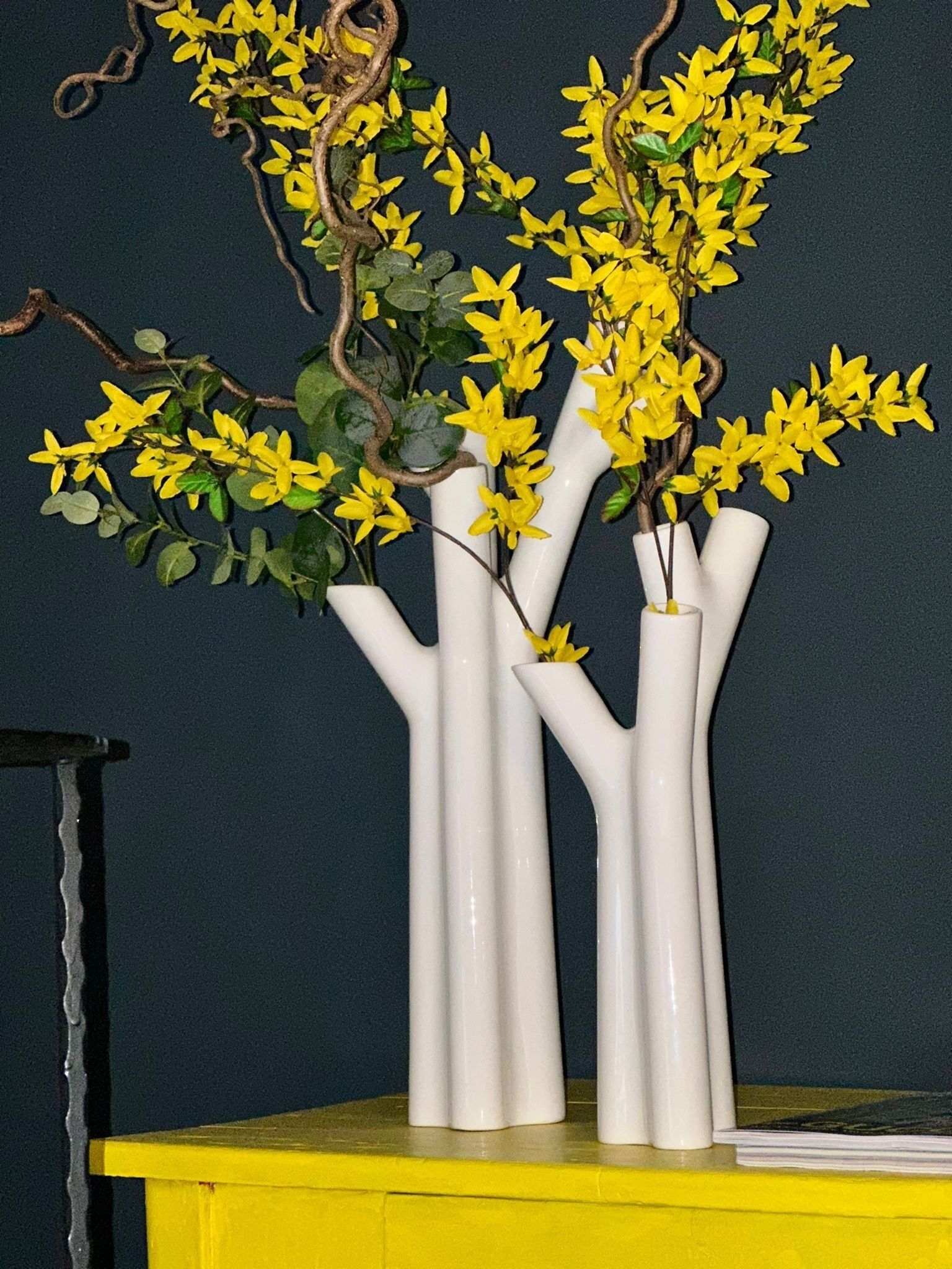 Tres jarrones blancos con flores amarillas sobre una superficie amarilla, sobre un fondo oscuro.
