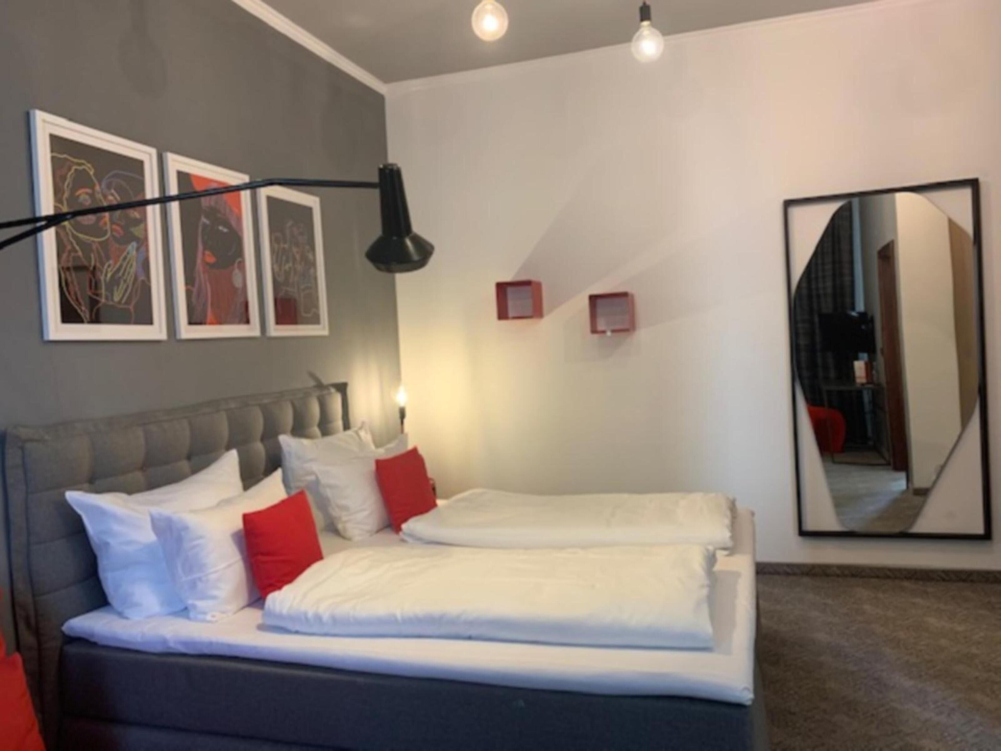 Een moderne slaapkamer met een grijs gestoffeerd hoofdeinde, wit beddengoed en rode accentkussens. Er staan twee nachtkastjes met lampen, en er hangt kunst aan de muren.