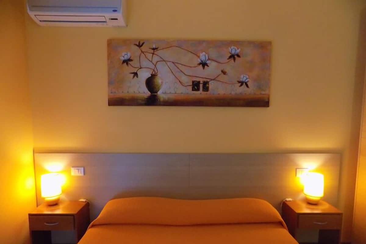 A hotel room with a bed, two bedside tables with lamps, an air conditioner, and a painting on the wall.