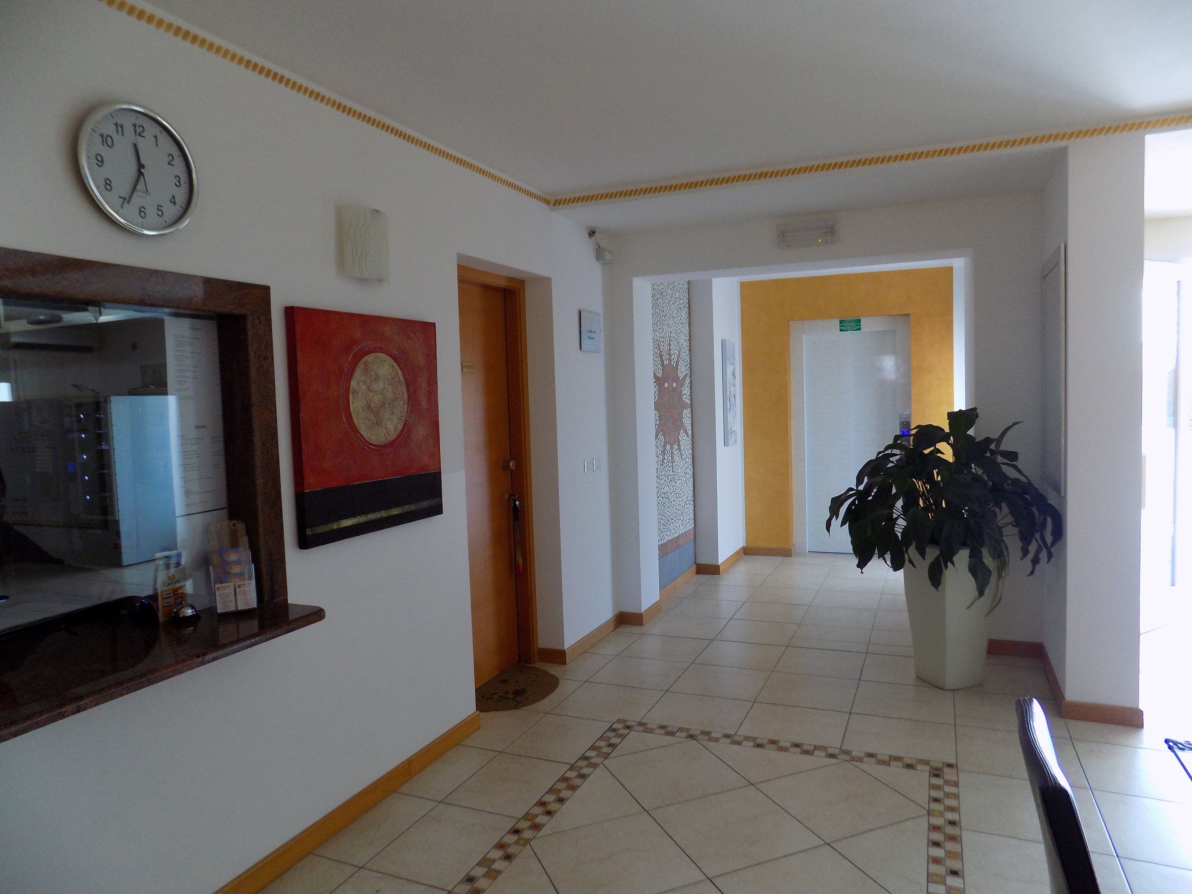 A hotel lobby with a clock, a reception desk, a large potted plant, and a patterned floor.