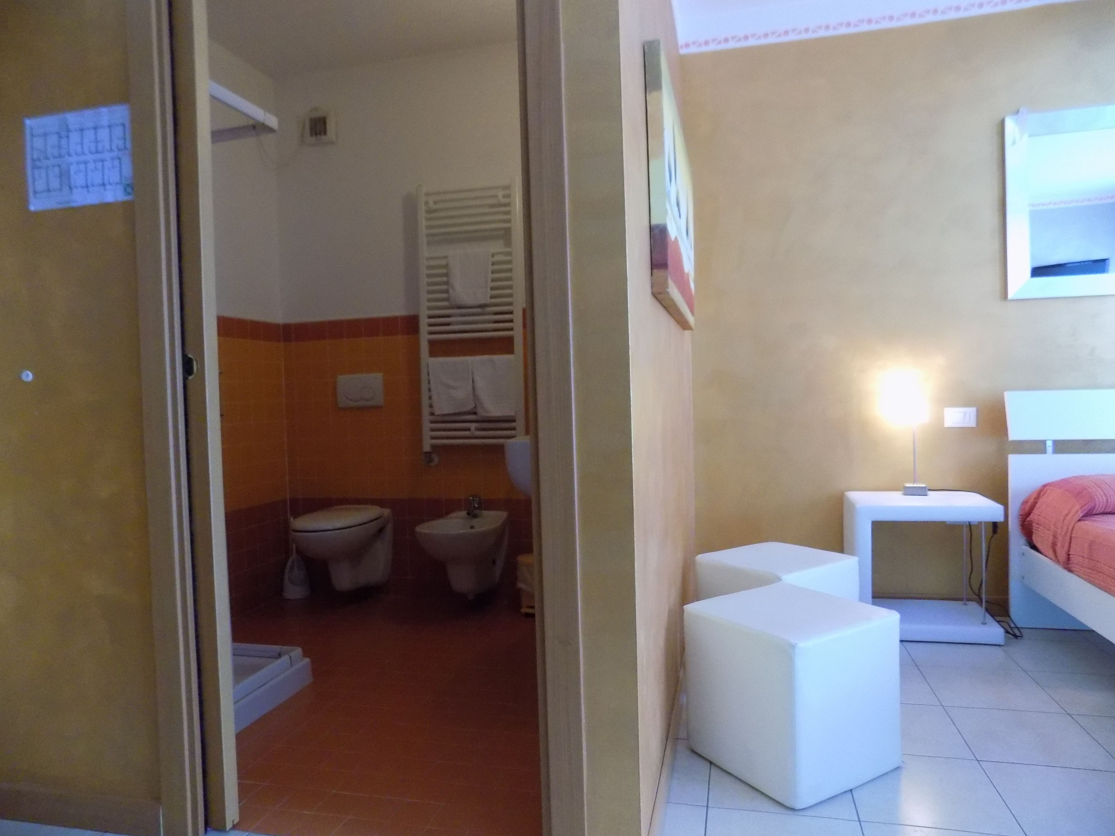 A small, compact room with a toilet, bidet, and sink. There is a bedside table with a lamp and a bed with a red blanket. The room has a tiled floor and a door leading to another area.
