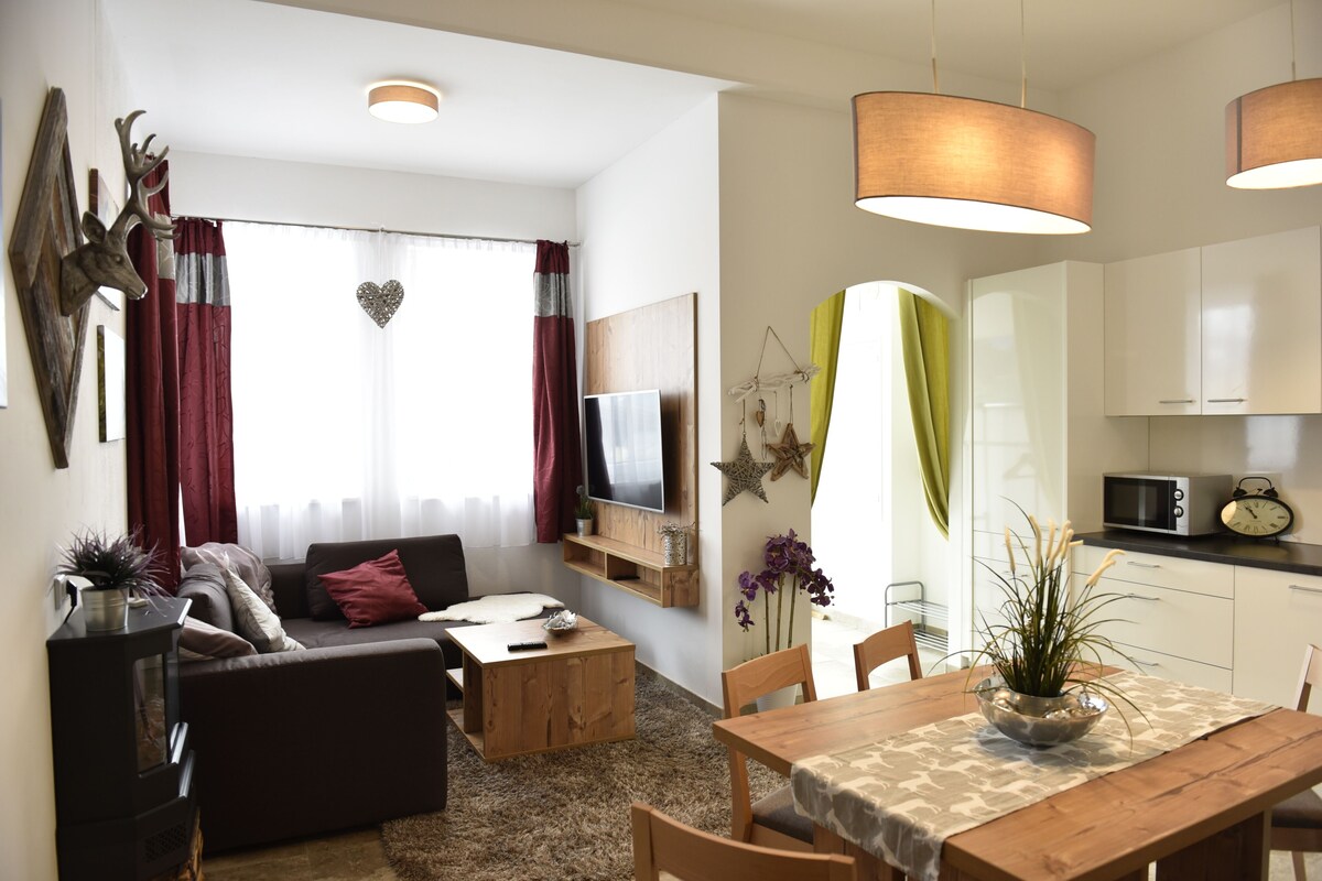 A modern living room with a sofa, coffee table, and TV. There's a dining area with a table and chairs. The kitchen has white cabinets and a microwave.
