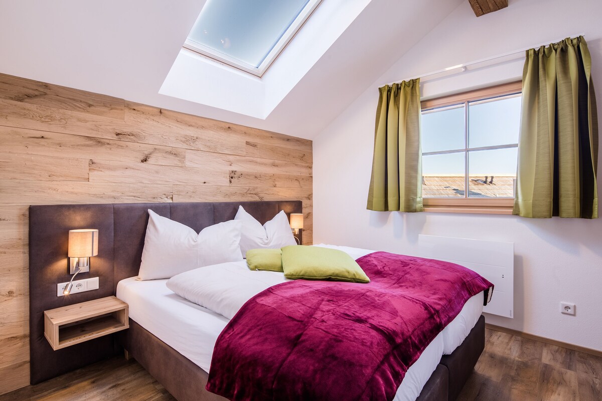 A bedroom with a skylight, a bed with white and green pillows, a red blanket, a nightstand with a lamp, and a window with curtains.