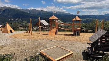 Ein Spielplatz mit Holzkonstruktionen, darunter eine Schaukel und Klettergerüste, vor einer Bergkulisse.