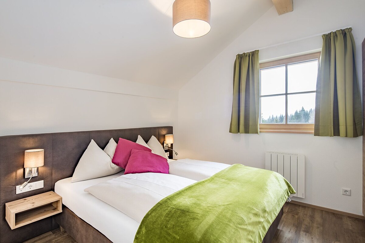 A neatly arranged hotel room with a large bed, a bedside table with a lamp, a window with curtains, and a radiator.