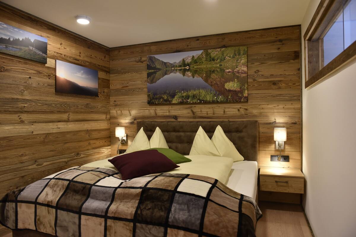 A wooden-paneled bedroom with a large bed, multiple pillows, bedside lamps, and framed landscape paintings on the walls.