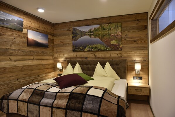 A wooden-paneled bedroom with a large bed, multiple pillows, bedside lamps, and framed landscape paintings on the walls. - Chaletdorf Fanningberg (Weisspriach)