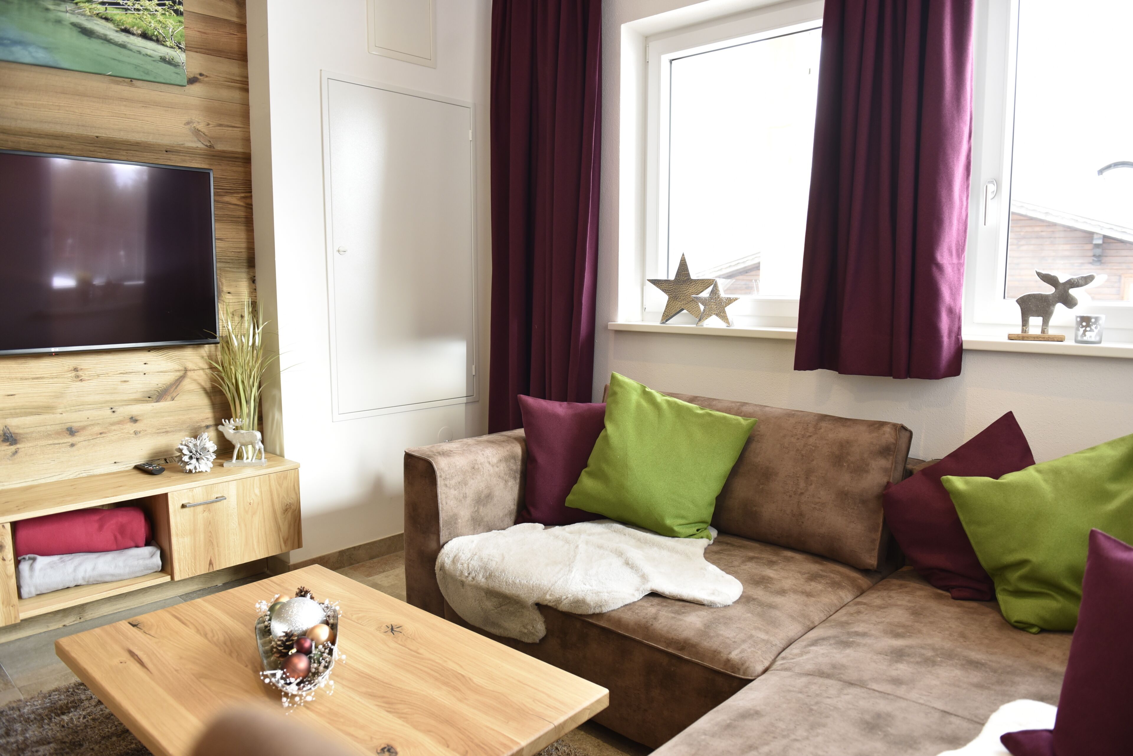 A living room with a brown sofa, green and maroon cushions, a wooden coffee table with a decorative item, a flat-screen TV on the wall, and a window with curtains.
