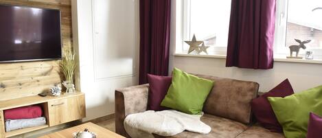 A living room with a brown sofa, green and maroon cushions, a wooden coffee table with a decorative item, a flat-screen TV on the wall, and a window with curtains.