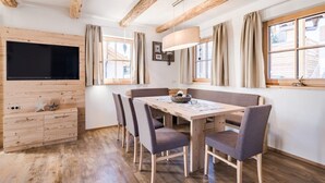 A dining area with a wooden table and chairs, a television mounted on a wooden wall, and a window with curtains. - Chaletdorf Fanningberg (Weisspriach)