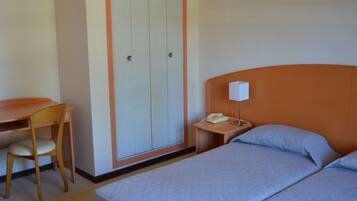 Una habitación de hotel con cama, escritorio, silla, armario y mesita de noche con lámpara y teléfono.