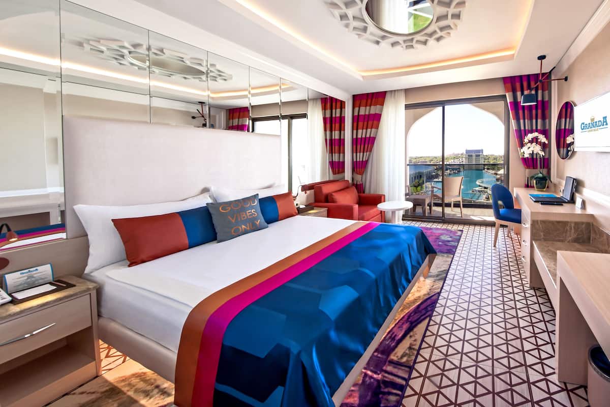 A modern hotel room with a large bed, a desk, a chair, a balcony with a view, and a large mirror.