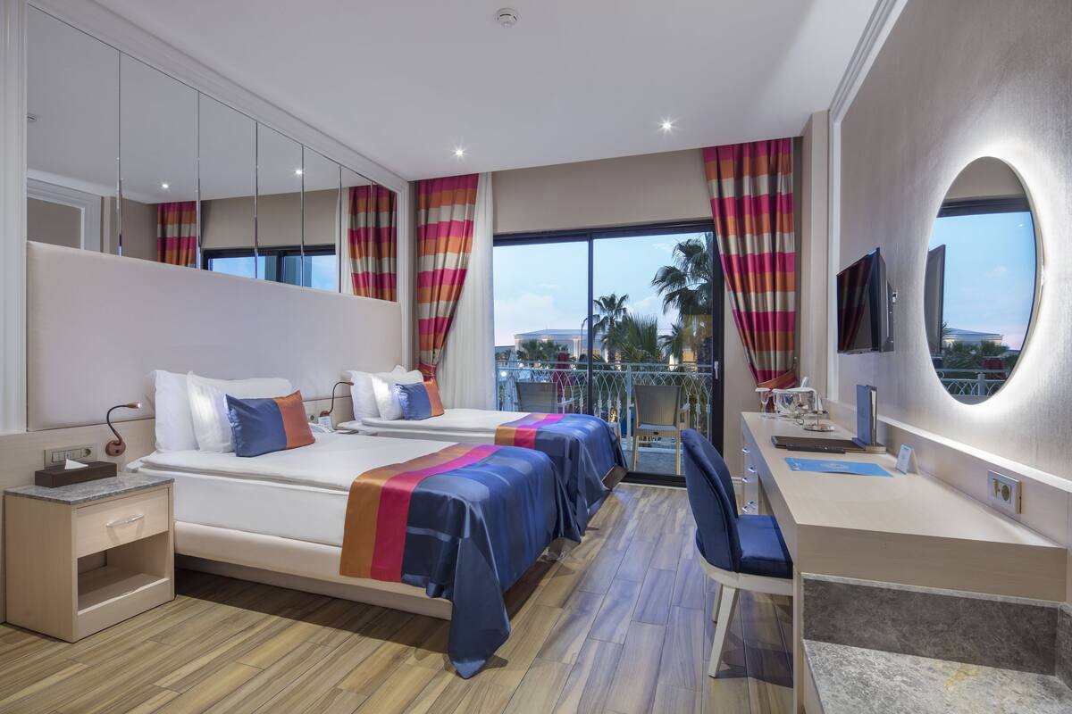 A modern hotel room with two beds, a desk, and a balcony with a view.
