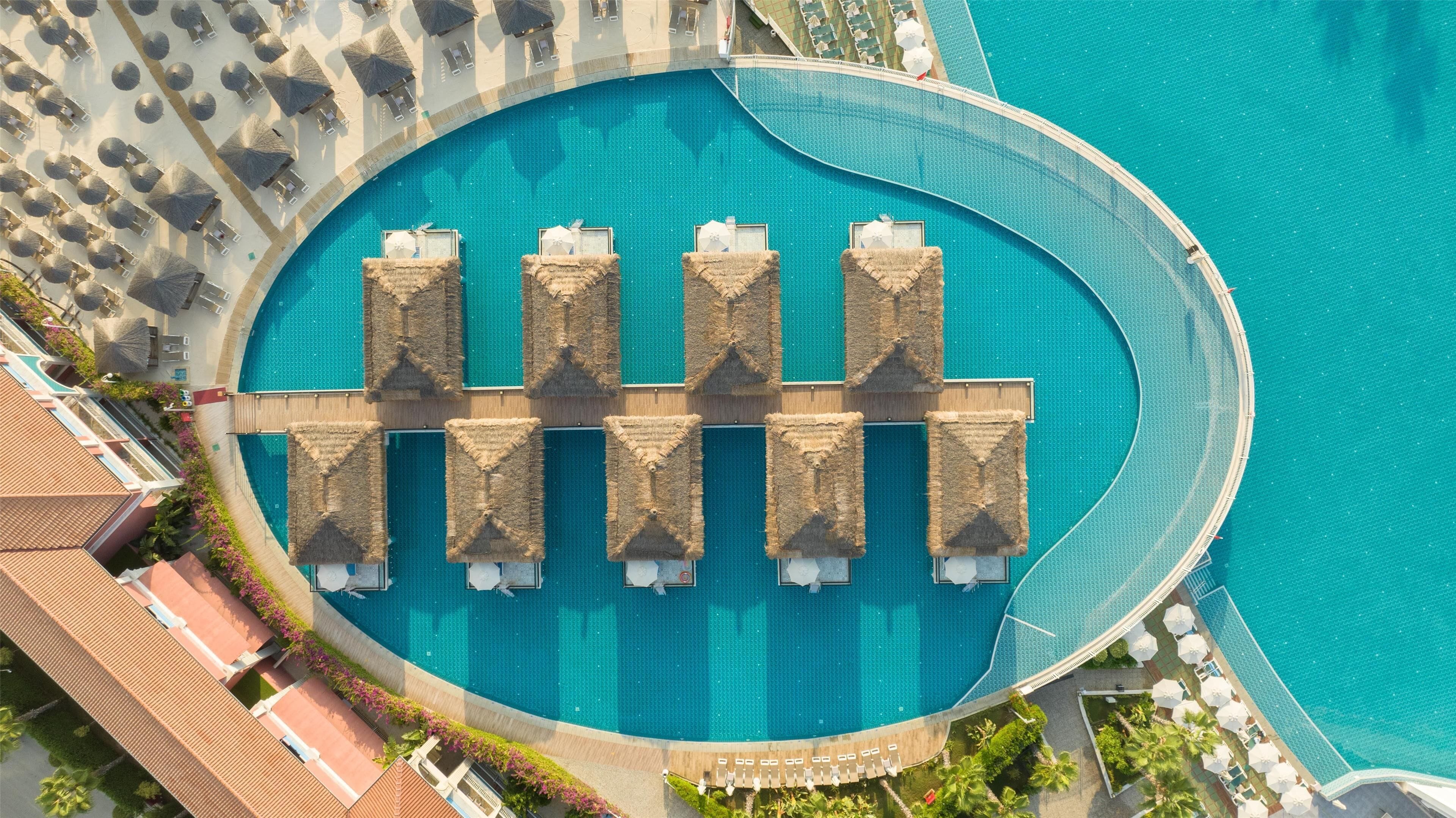 Eine Vogelperspektive auf einen kreisförmigen Pool mit Liegestühlen und Sonnenschirmen, umgeben von einer Poollandschaft mit einer Holzterrasse und Palmen.