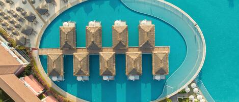Eine Vogelperspektive auf einen kreisförmigen Pool mit Liegestühlen und Sonnenschirmen, umgeben von einer Poollandschaft mit einer Holzterrasse und Palmen.