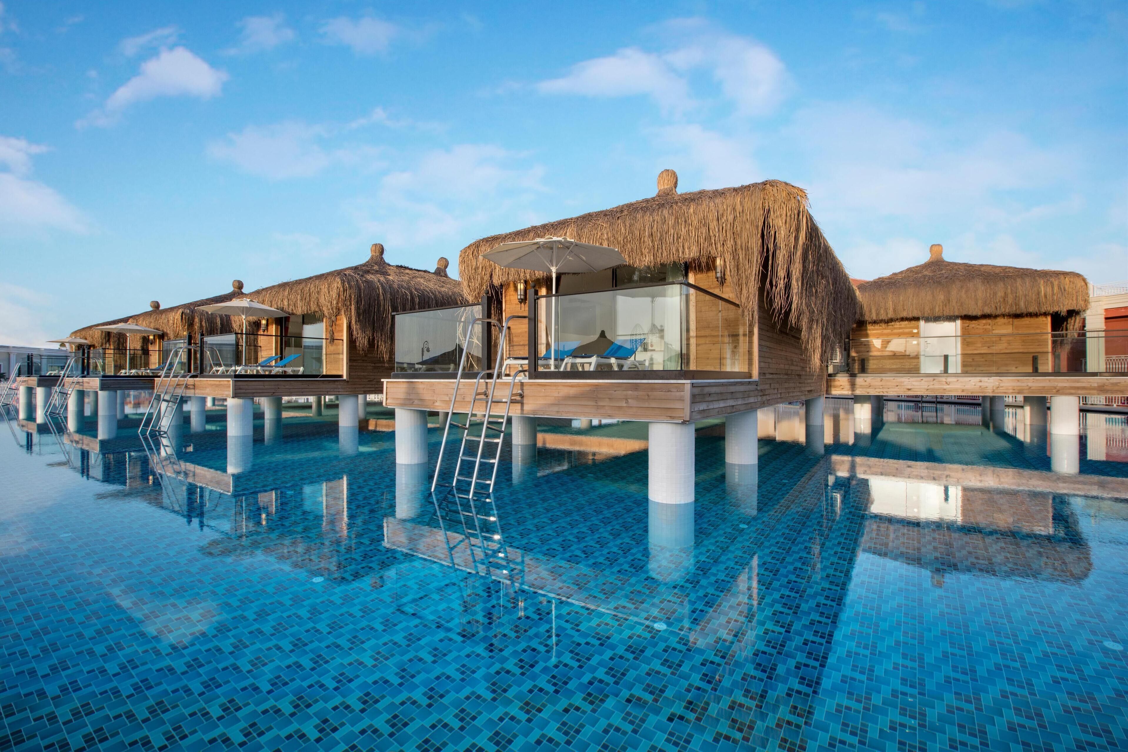 A poolside area with overwater bungalows, each with a thatched roof and glass windows.