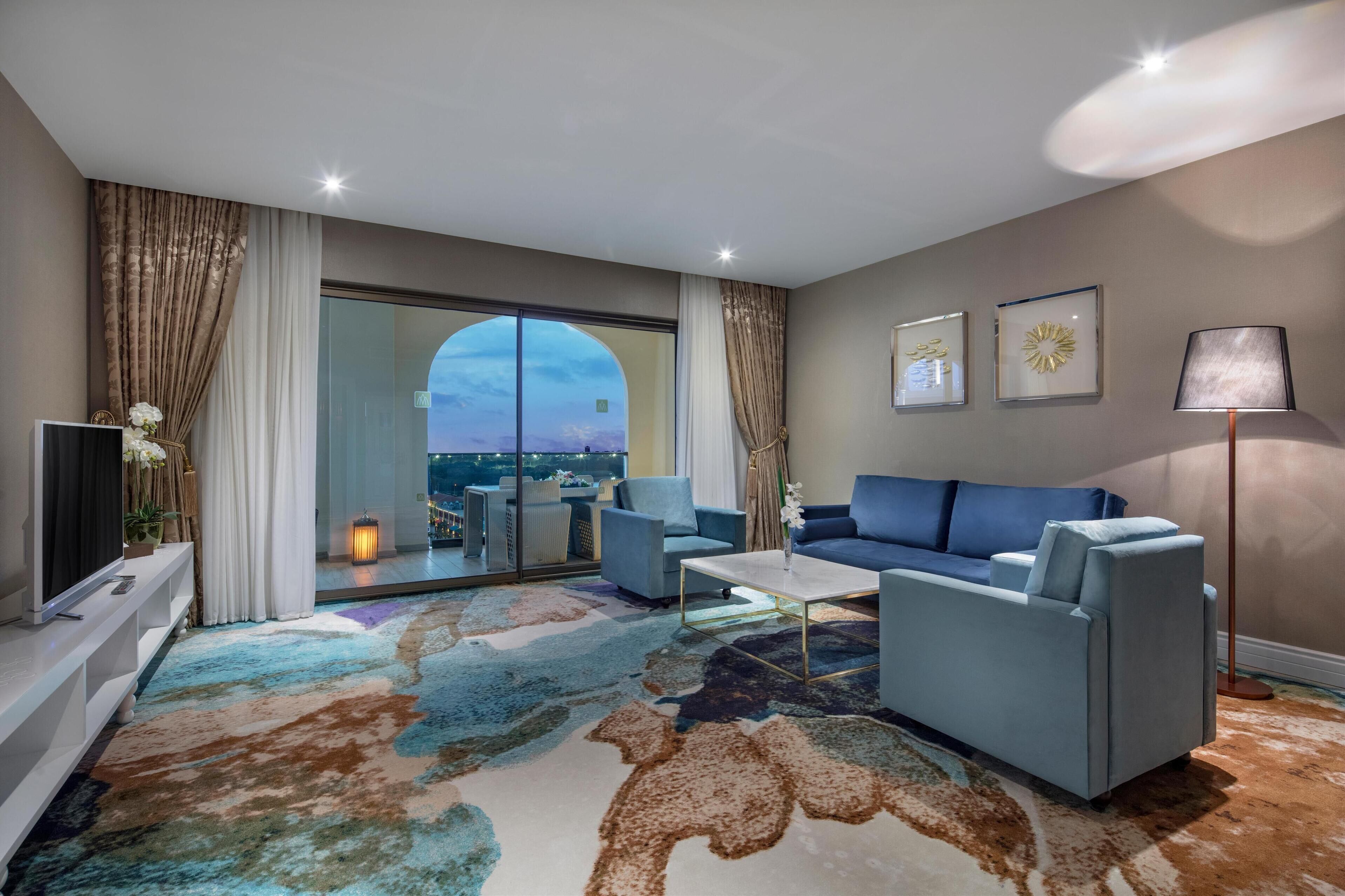 Una sala de estar moderna con un sofá, sillones, una mesa de centro y un televisor. Cuenta con un ventanal con cortinas, una lámpara de pie y dos cuadros en la pared.