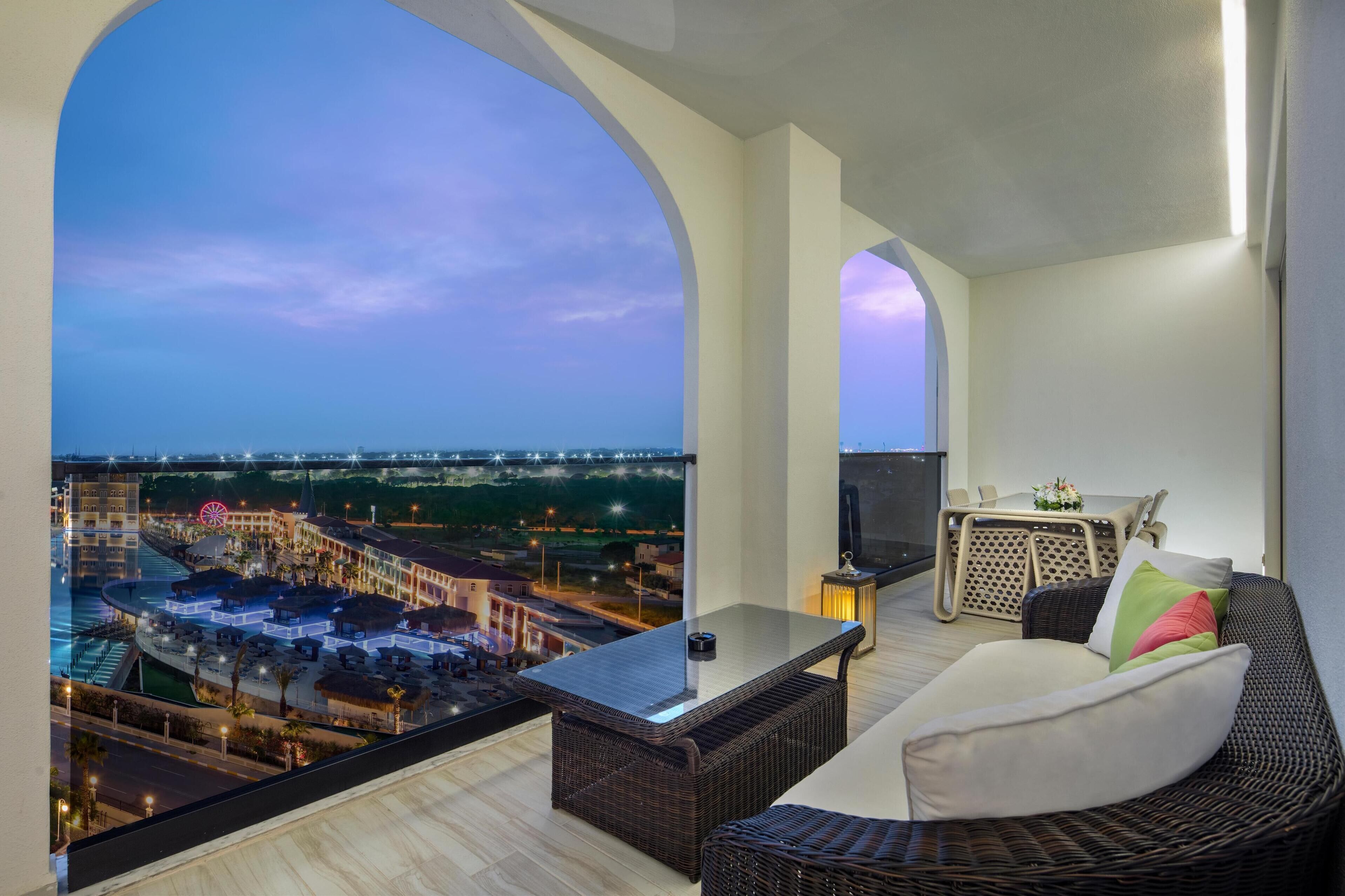 A balcony with a view of a cityscape at dusk, a wicker sofa, and a table with a TV.