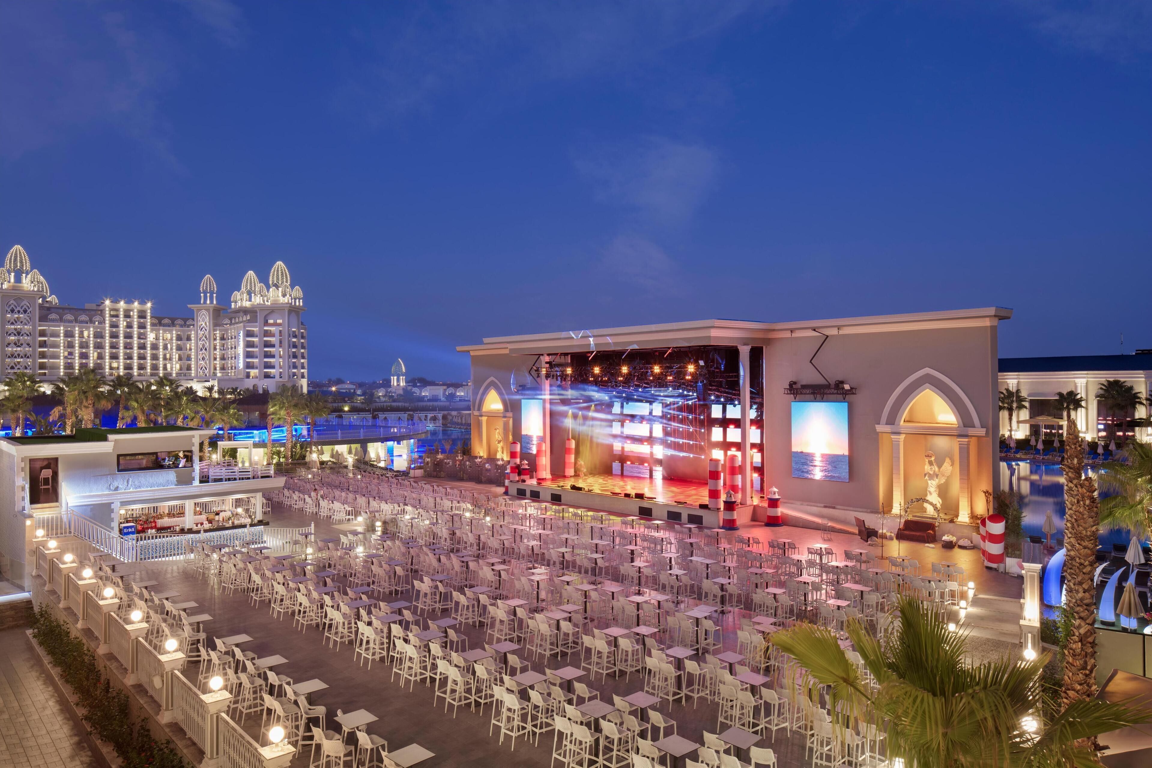 A rooftop dining area with tables and chairs, a stage with lighting, and a pool area.