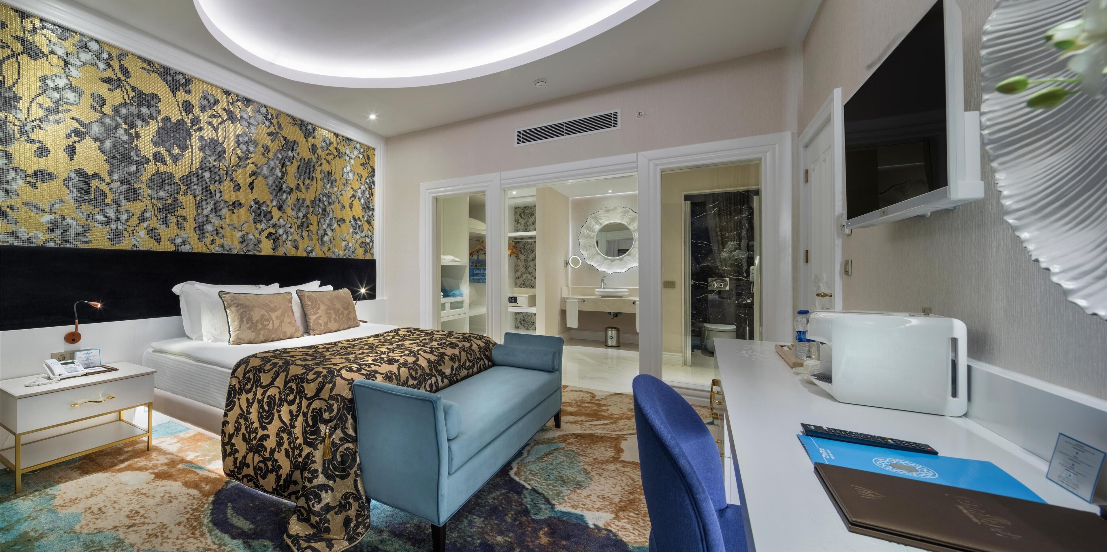 A modern hotel room with a bed, a desk, a chair, a nightstand, and a large mirror.