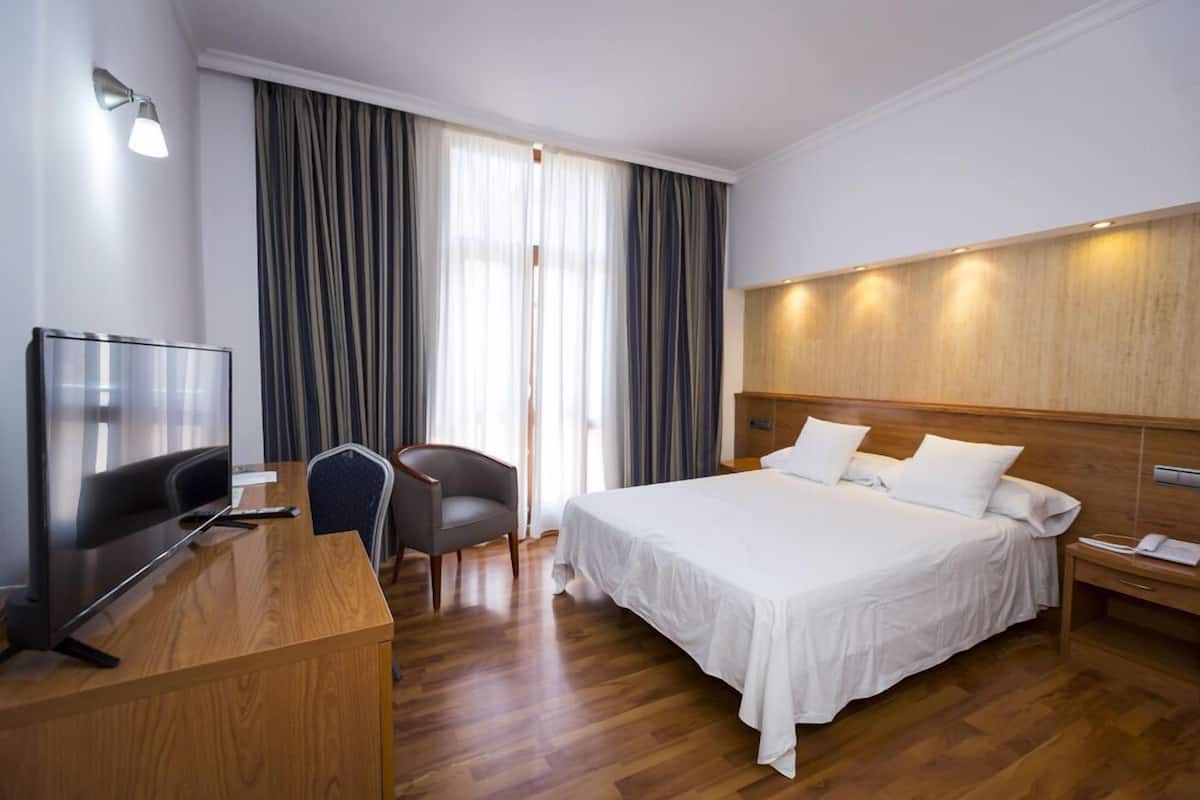 A hotel room with a wooden desk, a television, a bed with white linens, a chair, and a large window with curtains.