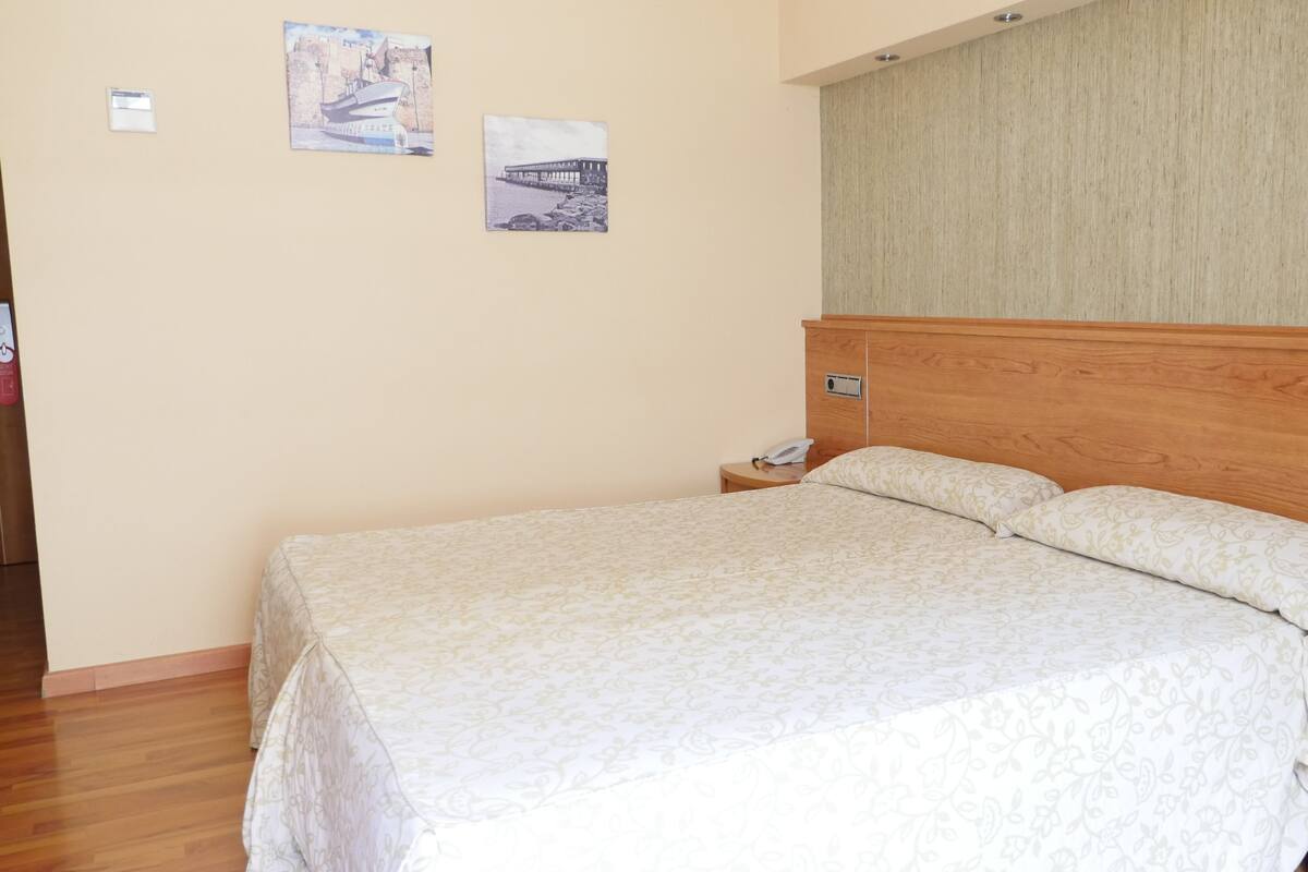 A neatly made bed with a wooden headboard, a bedside table with a phone, and two framed pictures on the wall.