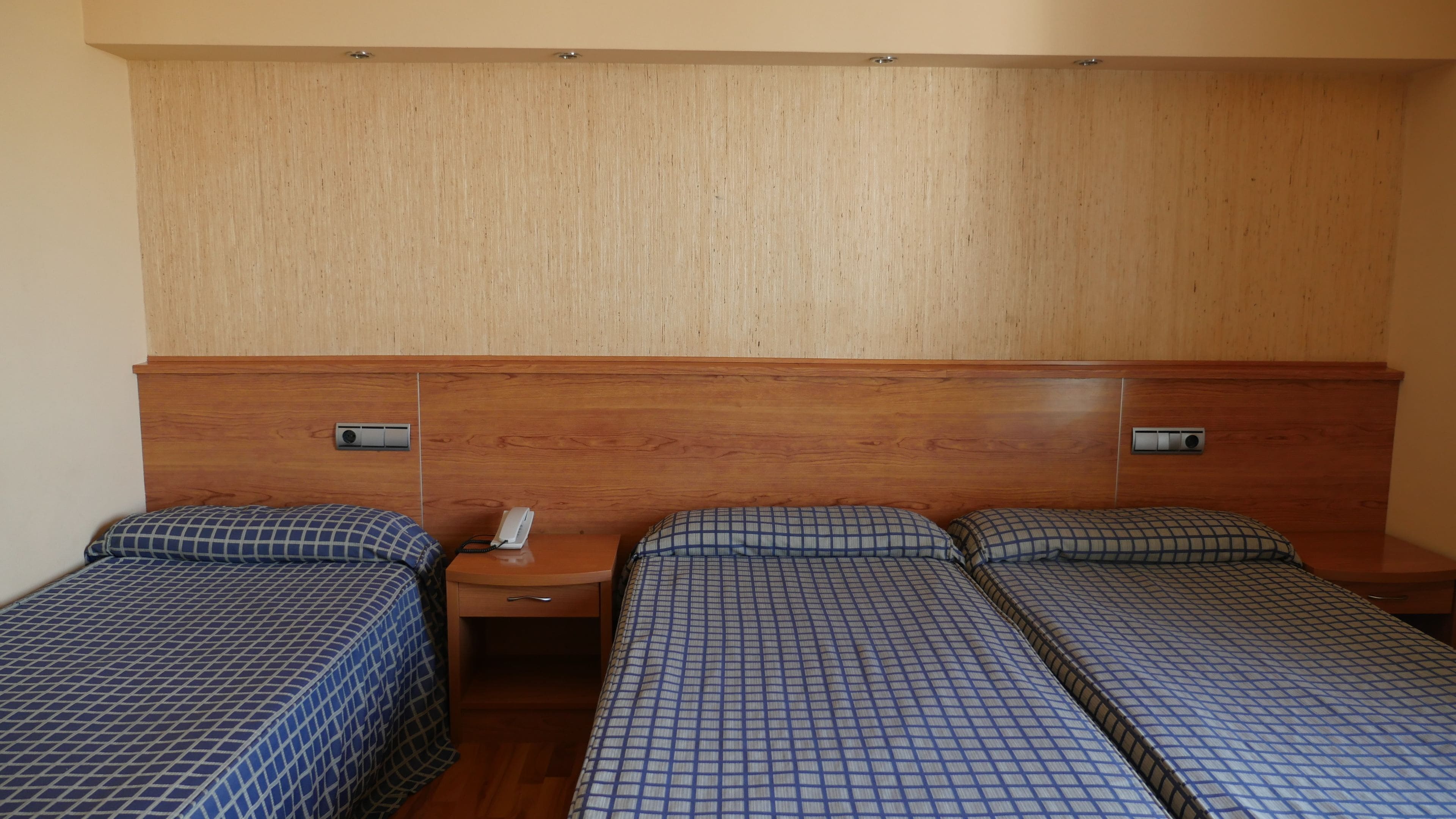 A hotel room with two single beds, a wooden headboard, a bedside table with a phone, and a wall-mounted air vent.