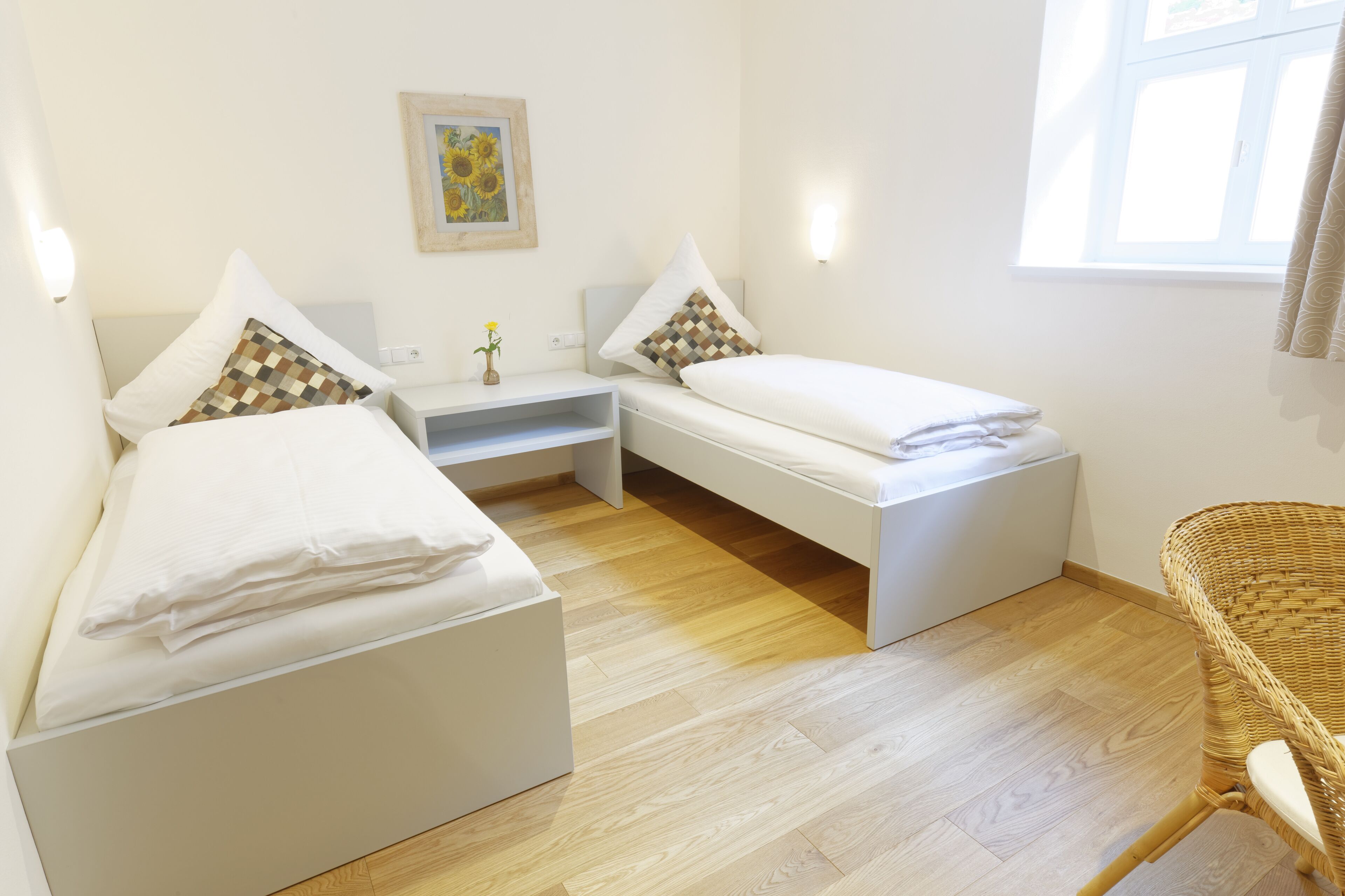 A twin room with two single beds, a small desk, a wicker chair, and a window with curtains.