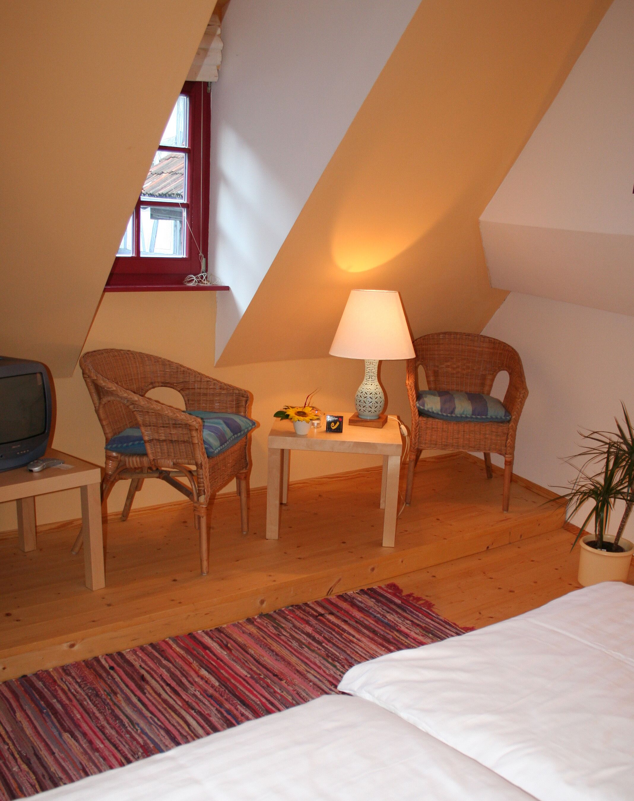 A cozy room with a bed, two wicker chairs, a small table with a lamp, a television, and a window with a red frame.