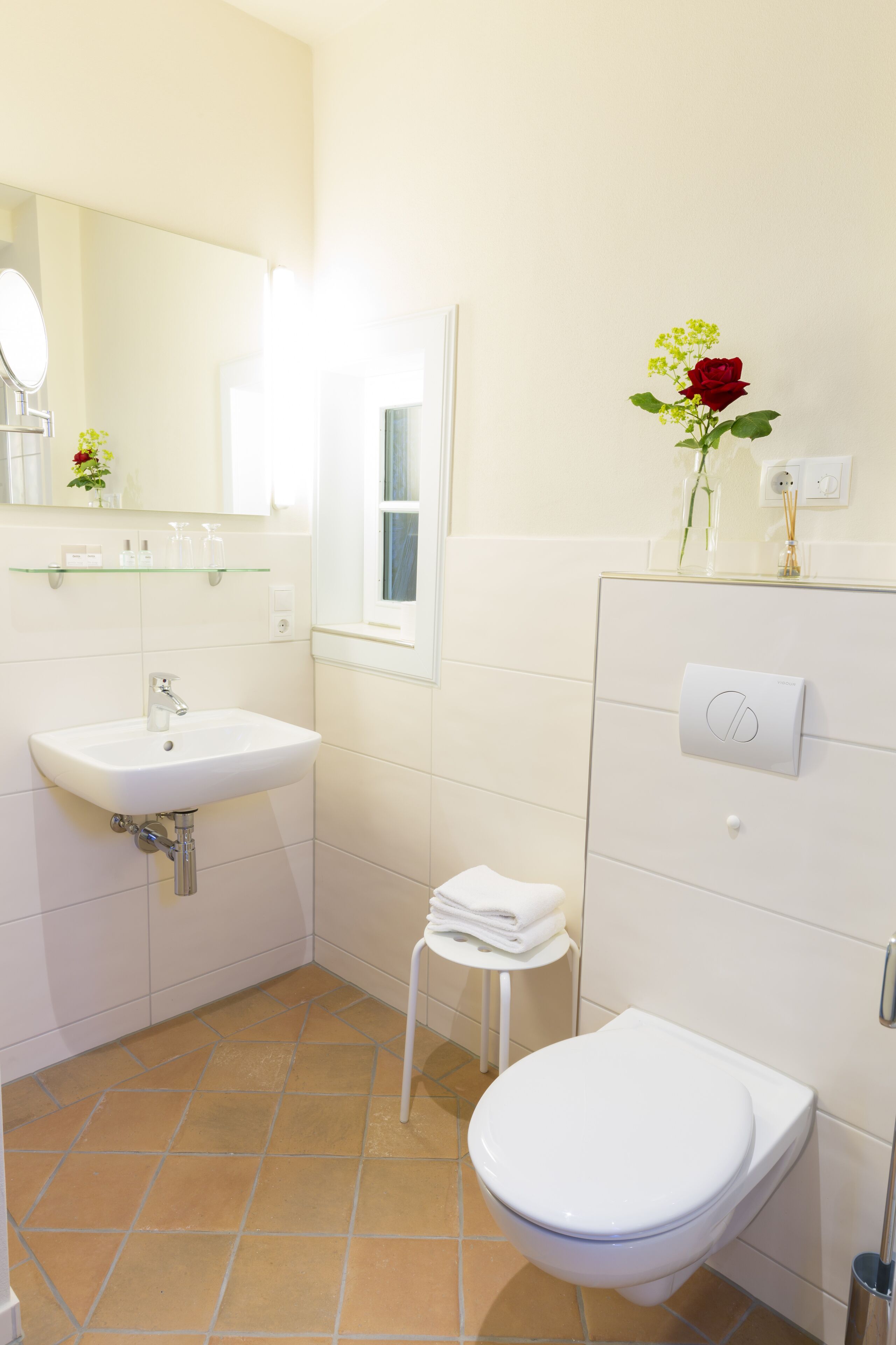 A bathroom with a white sink, mirror, and toilet. There is a small stool with a towel on it.