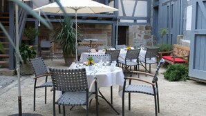 Zona pranzo all'aperto con tavoli apparecchiati per due, circondata da sedie e un ombrellone che offre ombra.