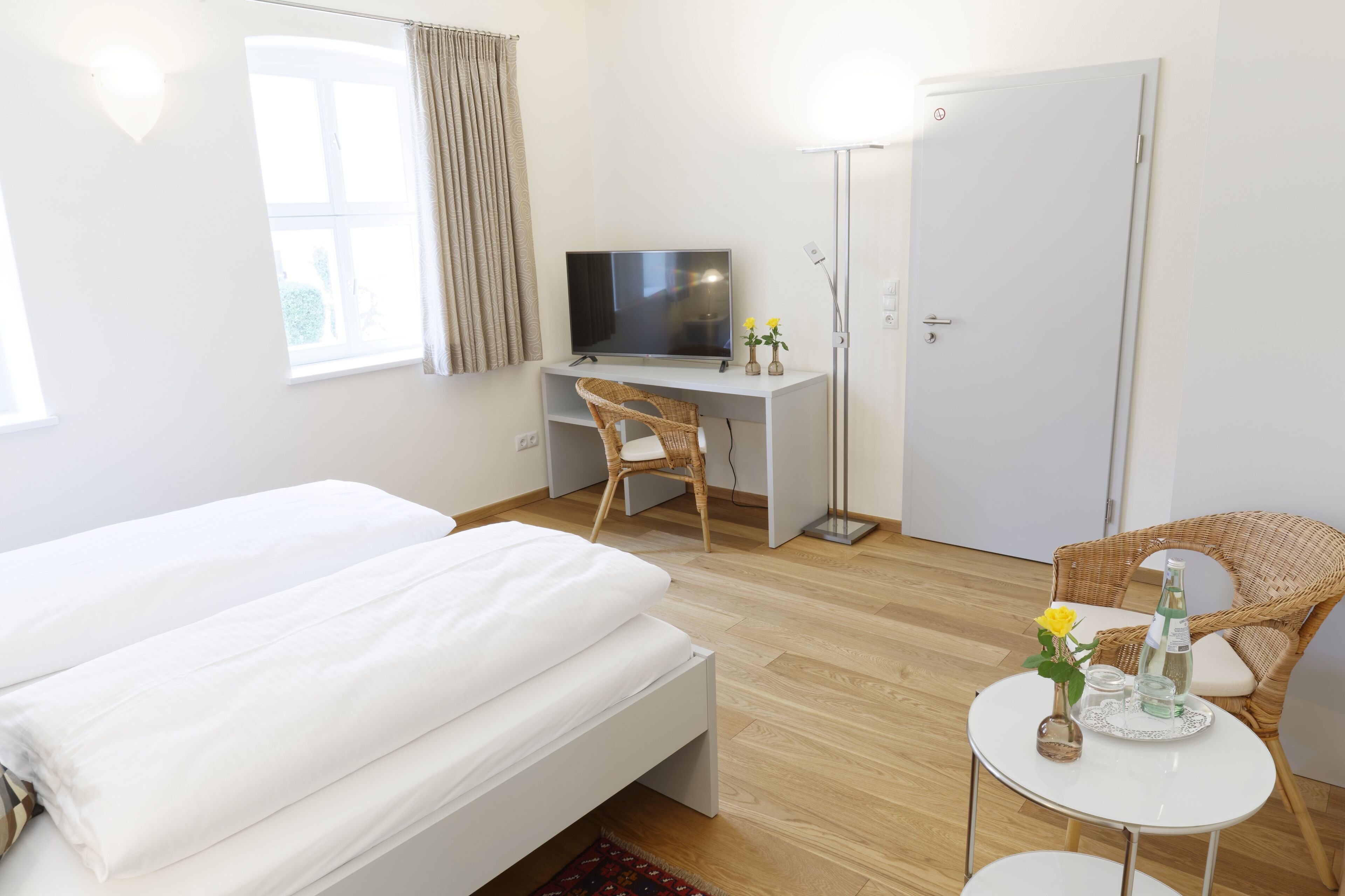 Habitación de hotel con cama, escritorio con televisor, silla, mesita con un florero y una botella, y una puerta.