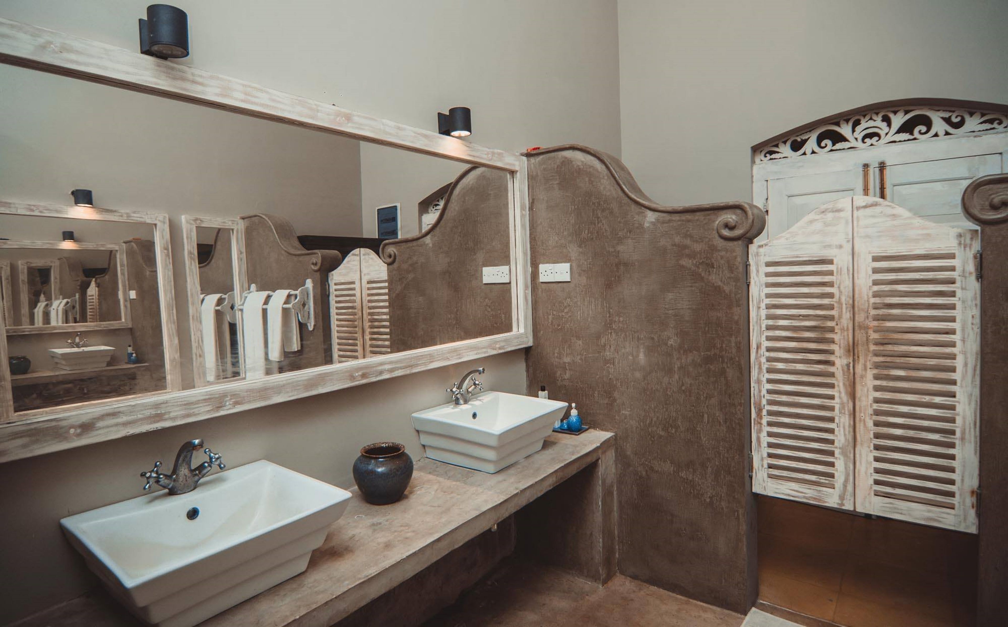 A bathroom with a large mirror, two sinks, a vase, and a wooden door with shutters.