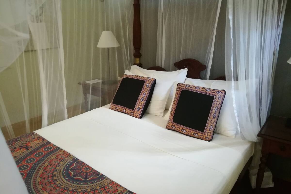 Ein Bett mit weißer Bettdecke, zwei Zierkissen, einem gemusterten Teppich und einem Baldachin mit transparenten Vorhängen.